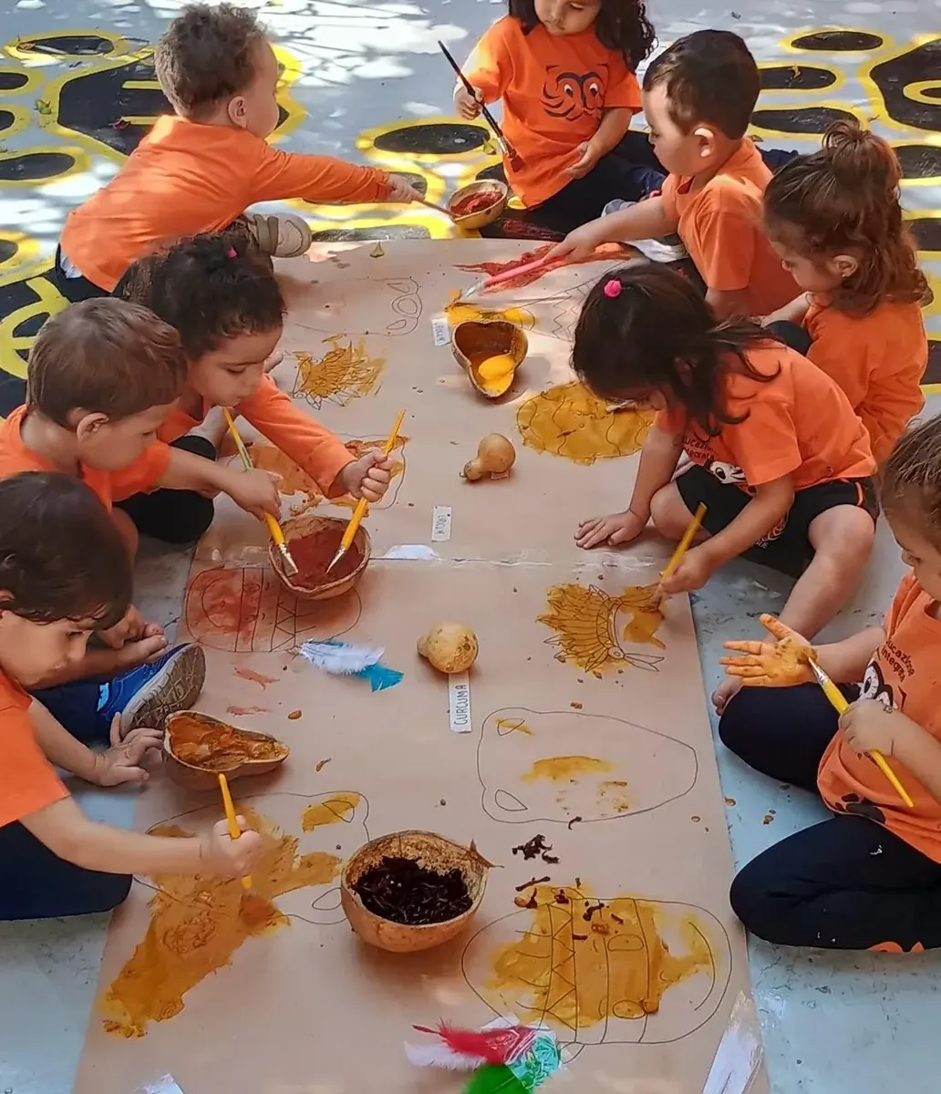 Crianças da Educação Infantil pintando um painel coletivo no chão do pátio, com pigmentos naturais em cuias