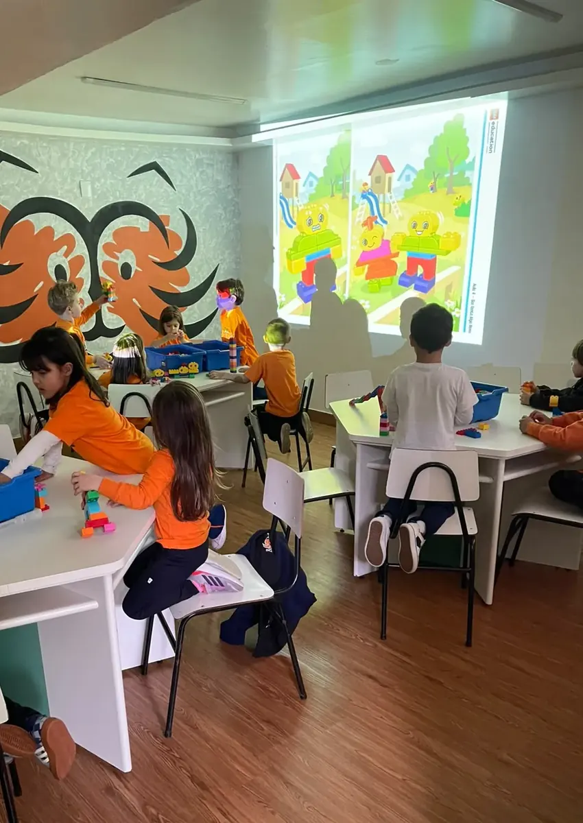 Turma da Educação Infantil na aula de Robótica, montando com blocos de encaixe grandes enquanto a atividade é projetada na parede