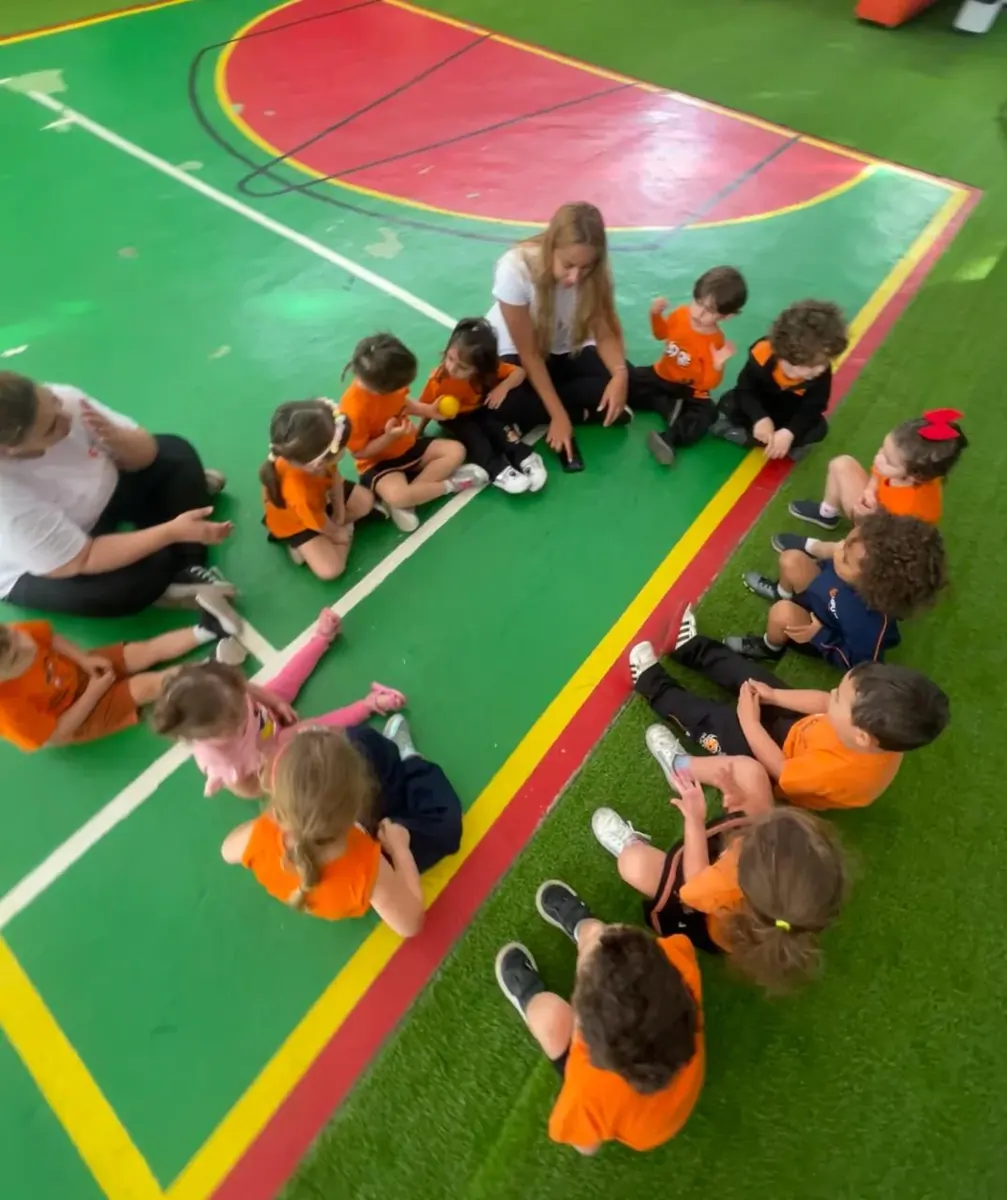 Turma da Educação Infantil sentada em roda na quadra do COLÉGIO HPLUS, com duas professoras